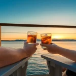 Couple holding up icy cocktail drinks while sitting on a seaside deck at sunset.
