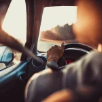 Uomo alla guida di un'auto, con le mani sul volante, guarda la strada davanti a sé, al tramonto.