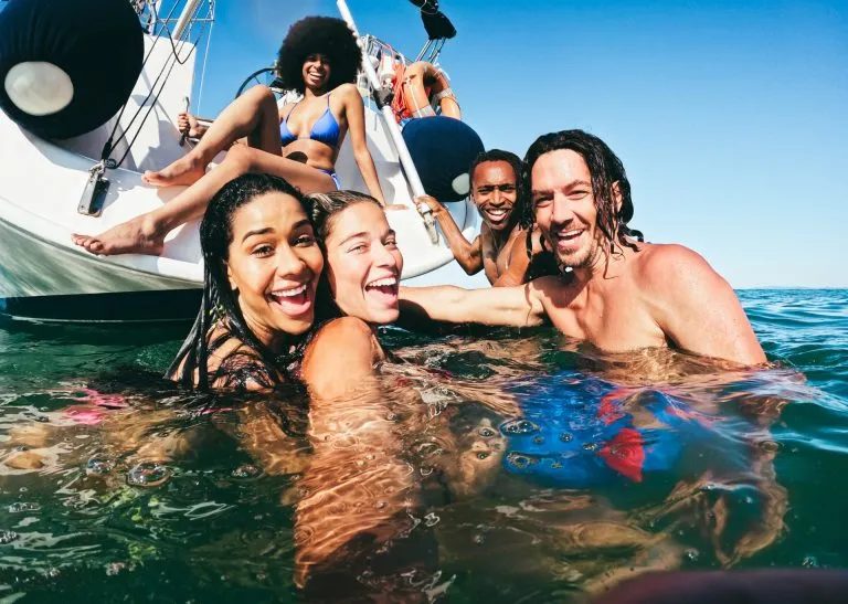Amigos multirraciales felices haciéndose un selfie nadando en el mar con un velero de fondo - Enfoque en el rostro del hombre de la derecha