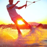 LENS FLARE, CLOSE UP: Young surfer wakeboarding and jumping 180 ollie at sunset